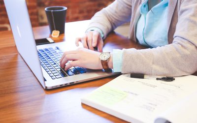 A close up of an individuals arms, stretched out working on a laptop alongside a mug of tea and a textbook under their left arm.