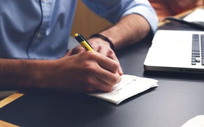 A close-up of an individuals arms and hands writing on a notepad and sitting at a desk in front of a laptop.