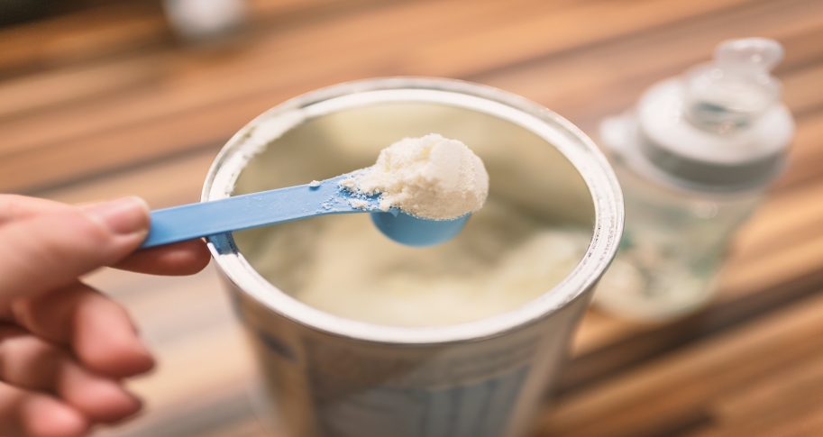 Powdered infant formula being scooped from a container on a kitchen table.