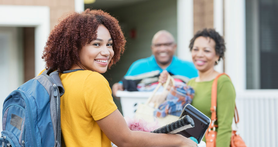 Parents helping their daughter relocate, perhaps into an apartment or college dorm. The young woman is in the foreground smiling at the camera, carrying a backpack and basket filled with her belongings.