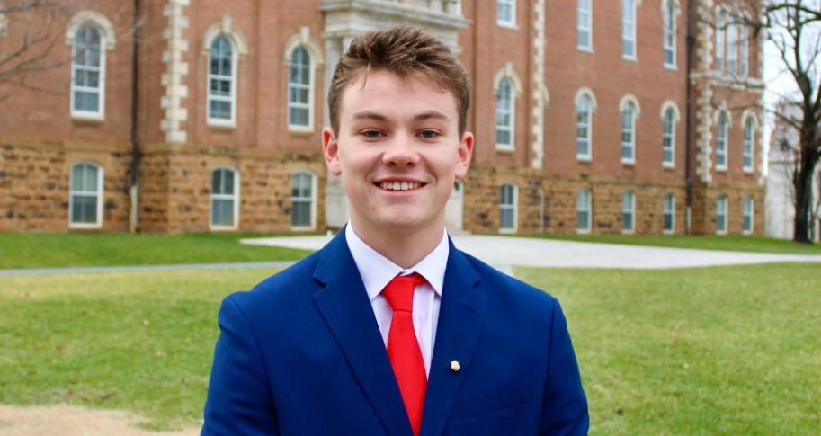 A student, Sam Butler in a blue suit and a red tie standing outside in from of Old Main.